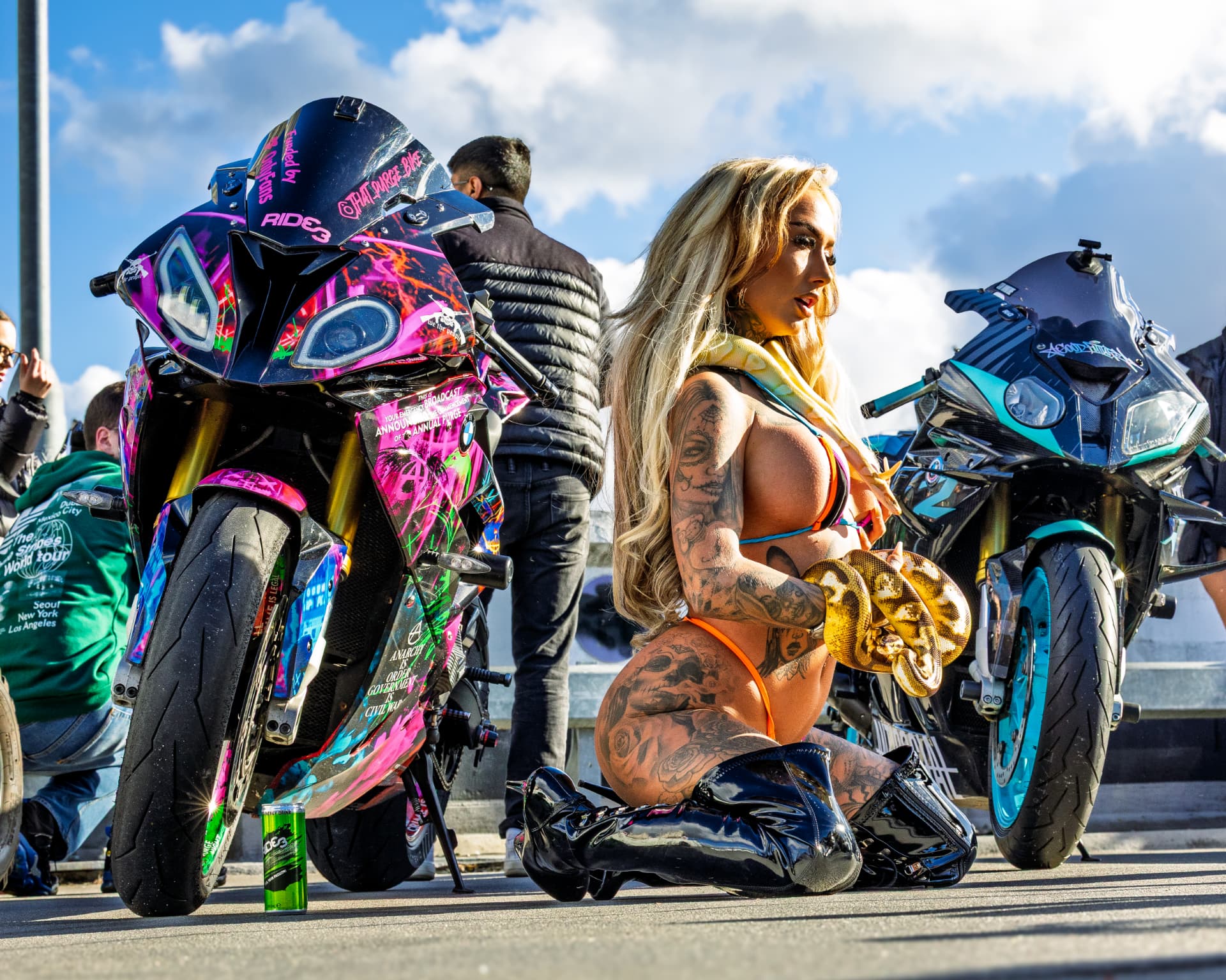 Model kneeling with a ball python between two custom-wrapped BMW superbikes at an outdoor event